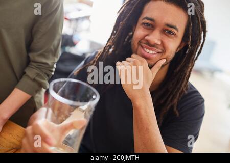 Afrikanischer Mann mit Dreadlocks sitzt lächelnd am Esstisch mit Freunden bei einer Mahlzeit Stockfoto