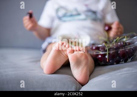 Süßer Kleinkind Junge, sitzt auf einer Couch, essen Kirschen, genießen gesunde Mahlzeit Stockfoto