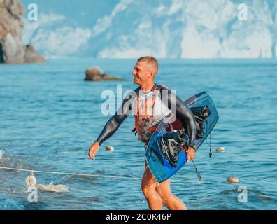 Glücklicher lächelnder Mann mit einem Brett für Wachen Surfen am Strand nach dem Reiten bei Sonnenuntergang. Wasserski. Ein männlicher Athlet spielt mit einem Brett Wassersport. Wake bo Stockfoto