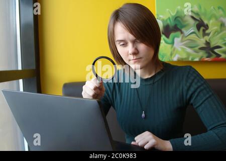 Ein junges, schönes Mädchen mit europäischem Auftritt in einer blauen Jacke, das in einem Café sitzt und auf einem Laptop auf der Couch vor dem Hintergrund einer gelben Wand arbeitet. Betrachtet ein Computerdisplay durch eine Lupe. Layout für Design und Ort für Text. Stockfoto