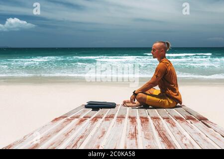 Junger Mann mit langen Haaren am Meer an einem sonnigen Tag, arbeitet mit seinem Laptop, arbeitet aus der Ferne mit einem Unternehmen. Bloner, Freiberufler, arbeitet durch das I Stockfoto