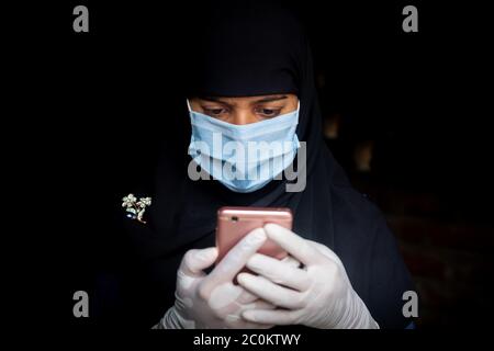 Eine Sicherheitsmaske trägt ein asiatisches muslimisches Mädchen ist mit seinem Smartphone. Schwarze Hijab Frau trägt eine blaue Maske für Coronavirus Sicherheit. Stockfoto