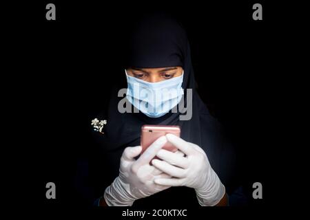 Eine Sicherheitsmaske trägt ein asiatisches muslimisches Mädchen ist mit seinem Smartphone. Schwarze Hijab Frau trägt eine blaue Maske für Coronavirus Sicherheit. Stockfoto