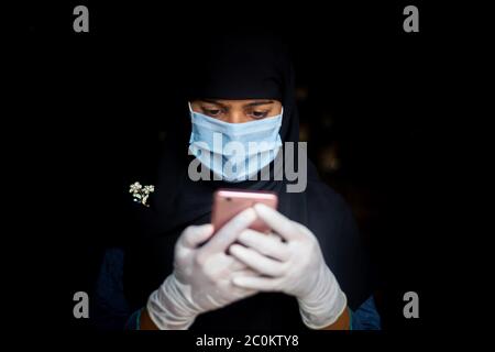 Eine Sicherheitsmaske trägt ein asiatisches muslimisches Mädchen ist mit seinem Smartphone. Schwarze Hijab Frau trägt eine blaue Maske für Coronavirus Sicherheit. Stockfoto