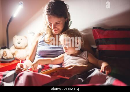 Mutter, ein Buch zu ihrem Kleinkind Jungen zu Hause in der Nacht zu lesen, Nachtstandlampe eingeschaltet, Mama und Baby umarmt Stockfoto