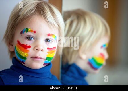 Schöne blonde Kleinkind Junge mit Regenbogen auf seinem Gesicht und unordentlichen Händen gemalt, lächelnd glücklich Stockfoto