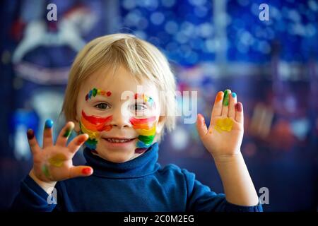 Schöne blonde Kleinkind Junge mit Regenbogen auf seinem Gesicht und unordentlichen Händen gemalt, lächelnd glücklich Stockfoto