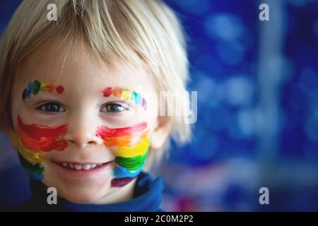 Schöne blonde Kleinkind Junge mit Regenbogen auf seinem Gesicht und unordentlichen Händen gemalt, lächelnd glücklich Stockfoto