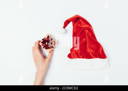 Frau Hand hält kleine Geschenkbox in der Nähe von roten Weihnachtsmann Hut auf weißem Tisch, Draufsicht. Stockfoto