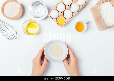 Weibliche Hände halten Schüssel mit trockener aktiver Hefe neben anderen Backzutaten auf weißem Küchentisch, Draufsicht. Stockfoto