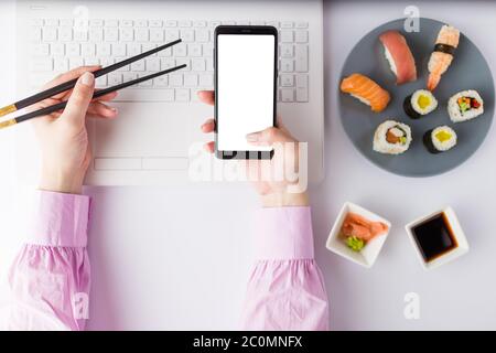 Flaches asiatisches Sushi-Set mit Laptop und weiblicher Hand, die Essstäbchen hält und leerem Handy. Gesunde Mahlzeit am Arbeitsplatz Lebensmittel-Delivery-Konzept. Stockfoto