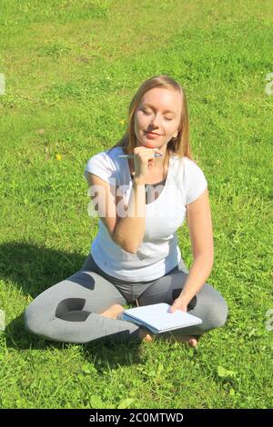 Ein junges, schönes Mädchen im weißen T-Shirt mit geschlossenen Augen sitzt auf dem grünen Gras auf dem Rasen mit einem Notizbuch und einem Stift, mit dem Handkinn gestützt, denkt und träumt, komponiert Text und Gedichte. Stockfoto