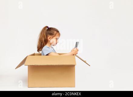 Ein kleines Mädchen sitzt in einem großen Karton und spielt auf dem Tablet. Stockfoto