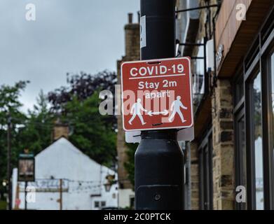 Ein rot-weißes Covid-19-Schild an einem Laternenpfosten vor den Geschäften in Baildon, Yorkshire. Das Schild weist Fußgänger an, 2 Meter voneinander entfernt zu bleiben. Stockfoto