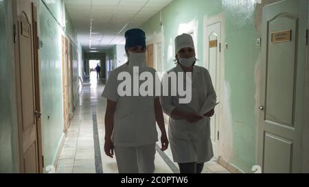 Zwei maskierte Krankenschwestern in weißen Mänteln, die durch den Krankenhaussaal gehen und die Kamera betrachten. Krankenhaus Innenraum im Prozess der Reparatur. Abstrakte Unschärfe-Unschärfe Stockfoto