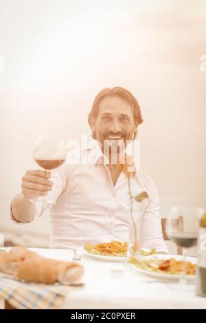 Lächelnd hält gut aussehend Glas Rotwein und schaut auf die Kamera, während er draußen am Tisch auf dem Meeresgrund sitzt. Festliches Toast-Konzept. Urlaub Stockfoto