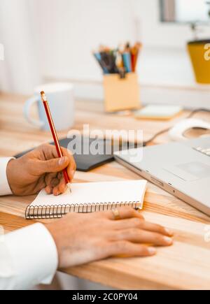 Creativeman macht Notizen in einem Notizblock. Nahaufnahme von dunkelhäutigen männlichen Händen mit rotem Bleistift und Schrift auf Papier Notizbuch. Getöntes Bild Stockfoto