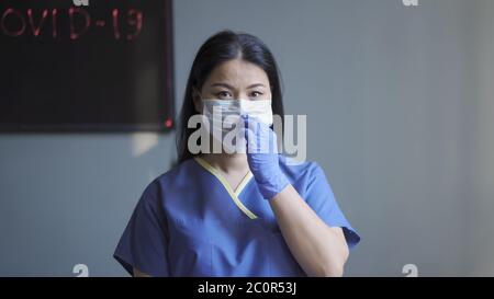 Maskierter Arzt, der in ihrem Büro arbeitet. Asiatische Frau in Maske und Schutzuniform Blick auf Kamera gegen graue Wand mit Neon Tafel und Stockfoto