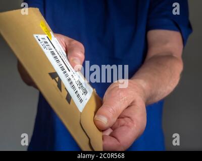 Mann in einem blauen T-Shirt liefert ein kleines Paket, indem er eine flache Pappverpackung mit Etikett vom Versand / Kurier / Lieferfirma übergibt. Stockfoto