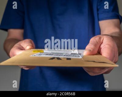 Mann in einem blauen T-Shirt liefert ein kleines Paket, indem er eine flache Pappverpackung mit Etikett vom Versand / Kurier / Lieferfirma übergibt. Stockfoto