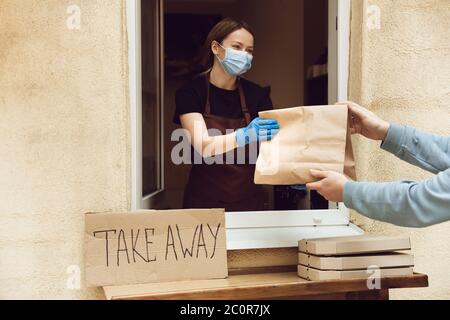 Frau, die Getränke und Mahlzeiten an den Kunden gibt, trägt eine Schutzmaske und Handschuhe. Kontaktloser Lieferservice während der Quarantänekoronavirus-Pandemie. Take Away-Konzept. Recycable Becher, Pakete. Stockfoto