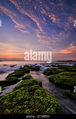 Schöner Sonnenaufgang in felsigen Strand mit grünem Moos bedeckt mit bunten Wolken am Himmel in Sawarna, Banten, Indonesien Stockfoto