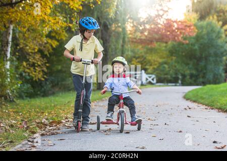 Schöne blonde zwei Jahre alten Kleinkind Junge und sein älterer Bruder, Reiten roten Dreirad und Roller im Park bei Sonnenuntergang, schönen Herbsttag Stockfoto