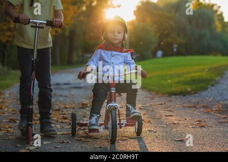 Schöne blonde zwei Jahre alten Kleinkind Junge und sein älterer Bruder, Reiten roten Dreirad und Roller im Park bei Sonnenuntergang, schönen Herbsttag Stockfoto