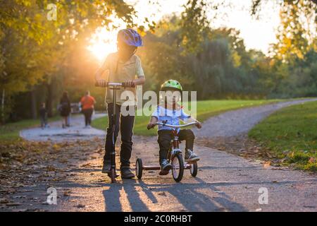 Schöne blonde zwei Jahre alten Kleinkind Junge und sein älterer Bruder, Reiten roten Dreirad und Roller im Park bei Sonnenuntergang, schönen Herbsttag Stockfoto