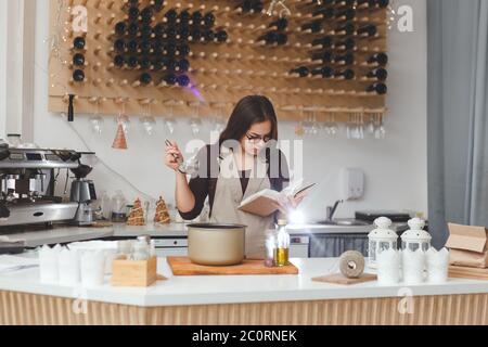 Junge Frau mit Schöpflöffel Kochbuch in der Küche lesen, auf der Suche nach Rezept der vegetarischen Suppe Stockfoto