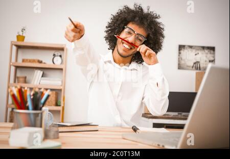 Fröhlicher Schüler, der Spaß während der Arbeitspause hat. Junger Araber im weißen Hemd lächelt breit und Gesten mit Bleistiften in den Händen. Getöntes Bild Stockfoto