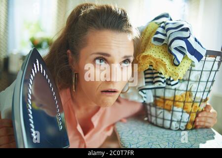 Portrait von traurig stilvolle Hausfrau in rosa Hemd und weiße Hose mit Eisen und gewaschen Wäschekorb bei modernen Hause in sonnigen Tag. Stockfoto