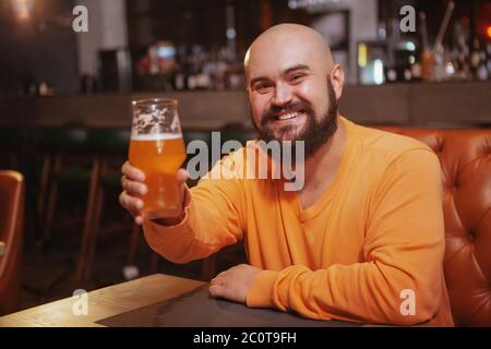 Fröhlicher, schöner bärtiger Mann, der fröhlich lächelt und mit seinem Bierglas im Pub toast Stockfoto