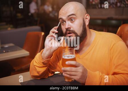 Ein hübscher Mann, der überrascht aussschaut und am Telefon telefoniert, während er im Restaurant Bier trinkt Stockfoto