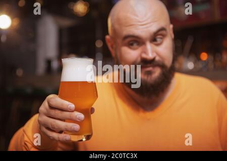 Nahaufnahme eines bärtigen, fröhlichen Mannes, der das Glas Bier in der Hand betrachtet und an der Bar trinkt Stockfoto