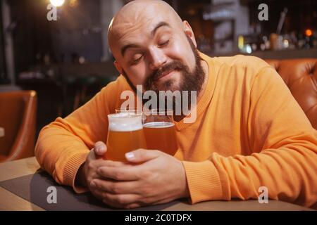 Charmanter junger Mann lächelt mit seinem geschlossenen Auge umarmende Bierbecher. Glücklicher Mann umarmte Biergläser Stockfoto