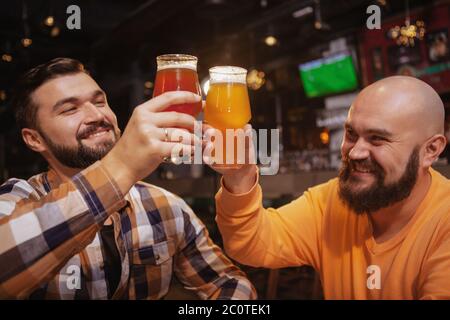 Glückliche männliche Freunde lachen, toasten mit ihren Bierkrügen. Gutaussehende bärtige Männer klirren Biergläser, feiern in der Kneipe Stockfoto