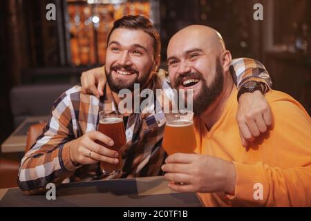 Glücklicher bärtiger Mann, der mit seinem besten Freund Bier trinkt. Männliche Freunde umarmen, halten ihre Biergläser hoch und feiern an der Bar Stockfoto
