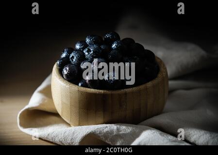 Frische Blaubeeren mit kleinen Tropfen Wasser auf der Oberfläche, in einer Bambusschale auf einem Holztisch, mit einer Tischdecke darunter. Stockfoto