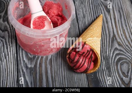 Erdbeereis in einem Waffelkegel. Garniert mit Schokolade. In der Nähe des Behälters mit Eis und einem Löffel dafür. Auf Pinien-Brettern in schwarz lackiert Stockfoto