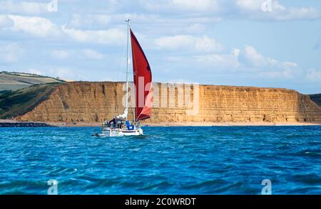 Eine Segelyacht passiert das Sandstein East Cliff, an der Jurassic Coast in der Nähe von West Bay, Dorset, UK. Berühmt gemacht durch die TV-Serie Broadchurch Stockfoto