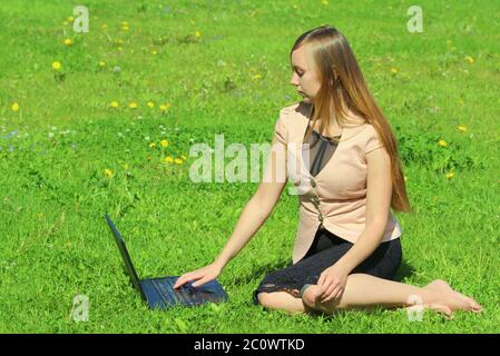 Ein schönes junges weißes Mädchen in einer rosa Jacke und schwarzem Rock und mit langen Haaren auf grünem Gras, auf dem Rasen sitzen und hinter einem schwarzen Laptop arbeiten. Stockfoto