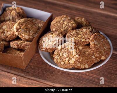 Handgemachte Haferflocken-Kekse auf einem weißen Teller und viele Kekse in einer Pappschachtel im Hintergrund auf einem Holz Stockfoto