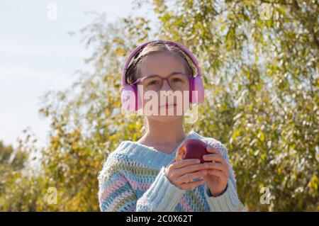 Schulmädchen mit Rucksack in Kopfhörer im Park. Ein Schulmädchen in rosa Kopfhörern hört Musik nach der Schule. Schulkindheit im Herbst. Hochformat o Stockfoto