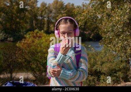 Schulmädchen mit Rucksack in Kopfhörer im Park. Ein Schulmädchen in rosa Kopfhörern hört Musik nach der Schule. Schulkindheit im Herbst. Hochformat o Stockfoto