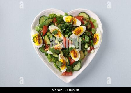 Herzförmiger Teller mit gesundem Salat mit Tomaten, Gurken, Eiern, Rucola und grünen Zwiebeln auf hellblauem Hintergrund, Blick von oben Stockfoto