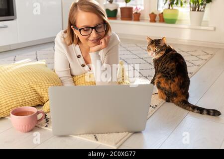 Freiberufliche Frau liegt im Wohnzimmer auf dem Teppich, Webinar ansehen. Frau mit ihrer Katze im Gespräch durch Video-Chat mit ihrer Familie, die in Isolation sind Dur Stockfoto