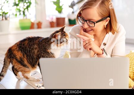 Lächelnde Frau Freiberufler liegt auf Teppich im Wohnzimmer, arbeitet auf Laptop zu Hause während der Selbstisolation, Katze in der Nähe will Aufmerksamkeit und Schlaganfall sein. Qu Stockfoto