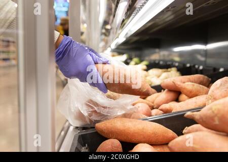 Frau trägt Gummi schützende medizinische Handschuhe, wählt Süßkartoffel Falten in Einweg-Plastiktüte im Supermarkt, aus der Nähe. Schutzmaßnahmen aga Stockfoto