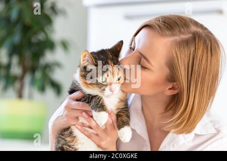 Nahaufnahme von lächelnden Frau in weißem Hemd küssen und umarmen mit Zärtlichkeit und Liebe Katze, halten sie in den Armen. Liebe zu den Tieren, Haustiere Konzept Stockfoto